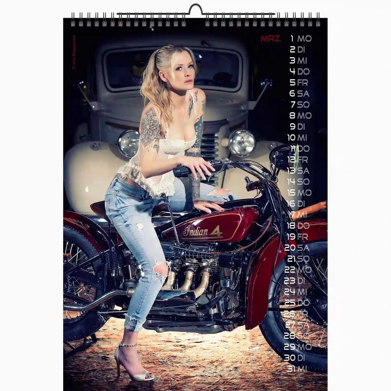Blonde tattooed woman sitting on a red vintage Indian motorcycle in a garage, with a classic car in the background, featured on the March page of the Bikes & Girls 2027 calendar.