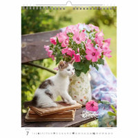 Kalenderpagina met een kat zittend op een houten oppervlak naast roze bloemen buitenshuis.